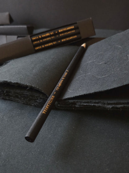 Black box of 6 eco friendly pencils, black in color. A person used a  newspaper pencil on the black deckled edge handmade cotton paper journal Black candy. White Canvaa India.