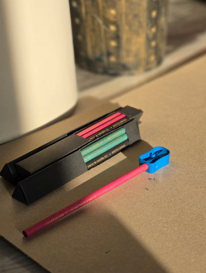 Two boxesof 6 eco friendly pencils each. Green, pink and black newspaper pencils Sharpening one pencil and sunlight is coming on it. White Canvaa India.